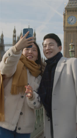 Video-Vertical-De-Una-Joven-Pareja-Asiática-De-Vacaciones-Posando-Para-Una-Selfie-Frente-A-Las-Casas-Del-Parlamento-En-Londres,-Reino-Unido